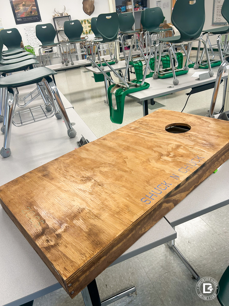 A finished stained cornhole board sits on a classroom table with “Shuck n’ Chuck” etched into the wood.