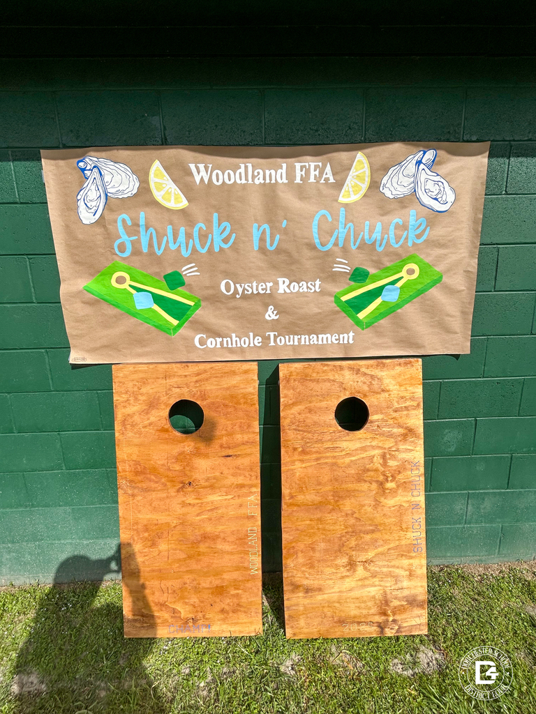 Two completed cornhole boards stand against a green wall beneath a sign that reads “Woodland FFA Shuck n’ Chuck Oyster Roast & Cornhole Tournament,” decorated with oysters, lemons, and cornhole boards.