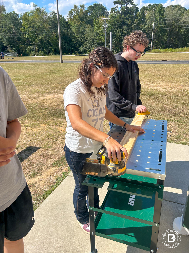 A student sands a wooden board with a power sander on an outdoor workbench while another student watches.