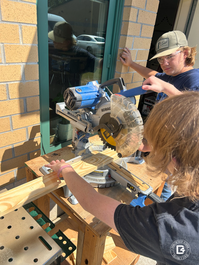 Two students use a miter saw outside the classroom to cut a piece of wood while wearing safety glasses.