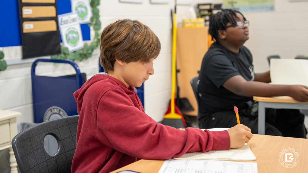 Two students work quietly at their desks, one writing in a notebook and another reading a paper during class.