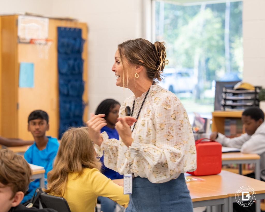 Mrs. Ayer enthusiastically gestures while leading a discussion with students seated around the classroom.