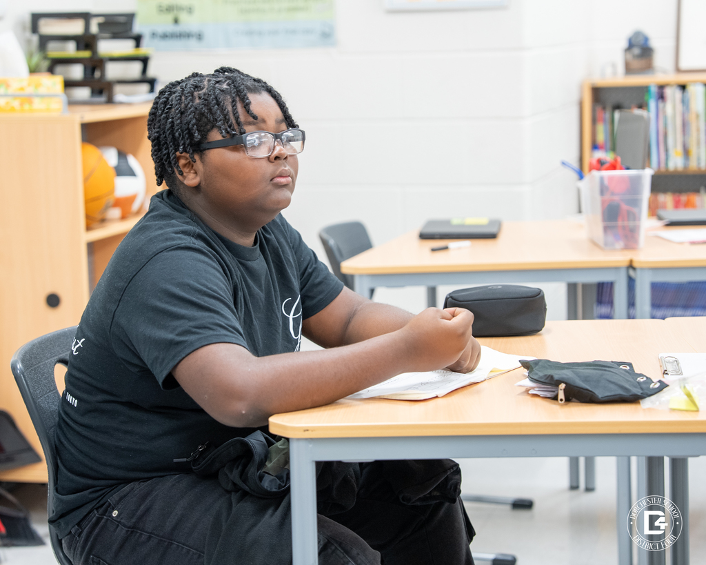 A student wearing glasses listens carefully during an English Language Arts lesson, with papers spread on his desk.