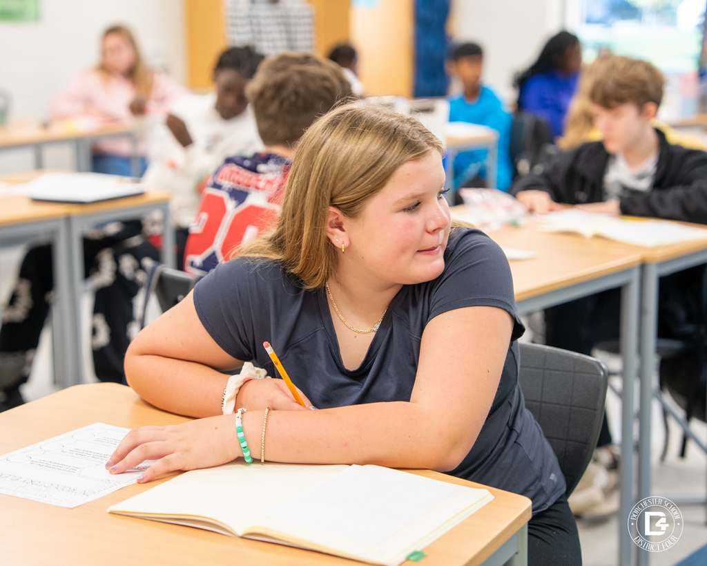 A student smiles slightly while looking across the classroom, holding a pencil and notebook on her desk.