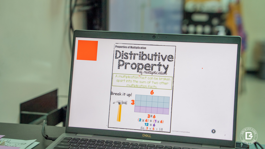 A laptop displays a colorful slide about the distributive property of multiplication, showing an example problem broken into smaller parts using visuals and numbers.