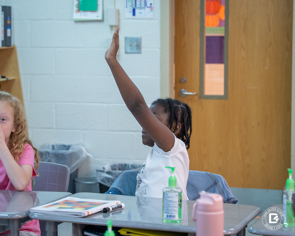 A student raises her hand eagerly to answer a question while another student watches attentively during a math lesson on multiplication.