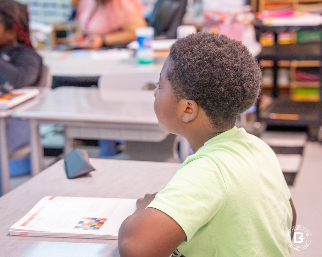 A student in a light green shirt listens attentively during the math lesson, with a workbook open on the desk.
