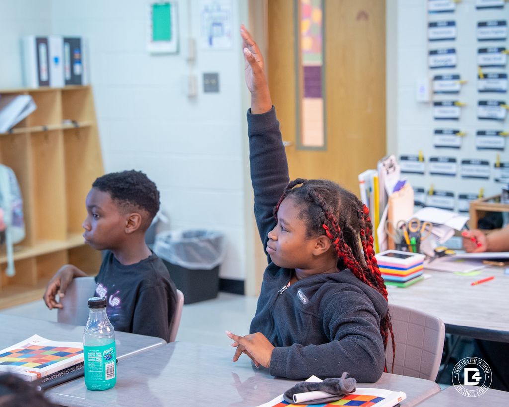 A student with red braids raises her hand confidently to participate in a classroom discussion about multiplication while another student listens beside her.