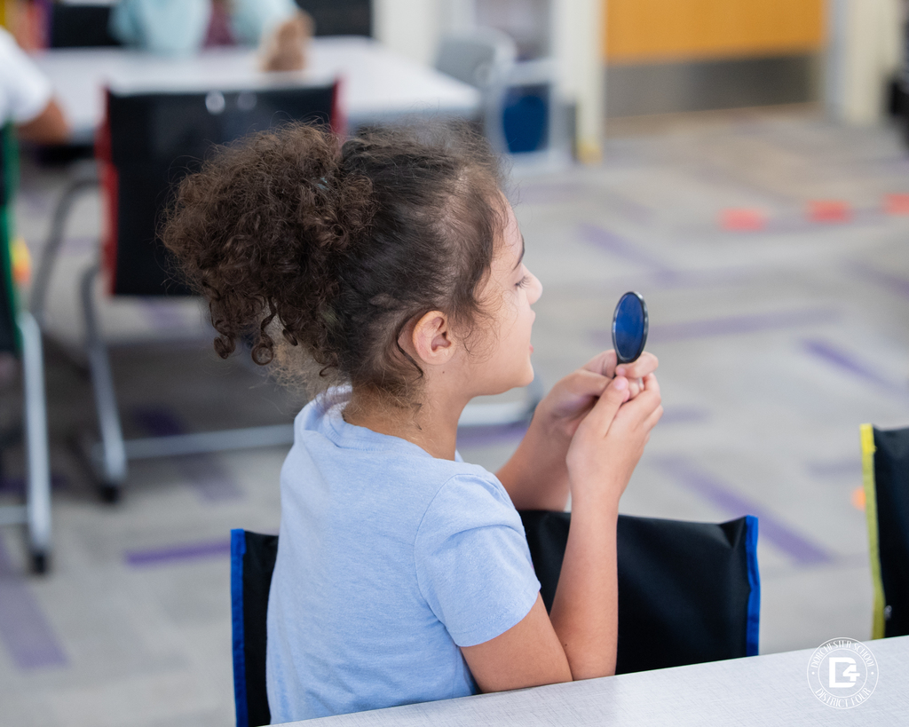 A student in a blue shirt uses a handheld mirror to observe how their lips and tongue move while making different letter sounds.