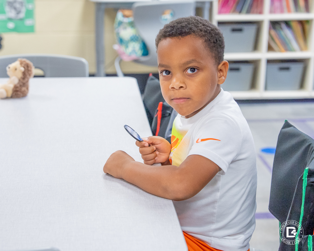 A student in a white shirt holds a mirror and looks toward the camera during a literacy lesson focused on sound recognition.