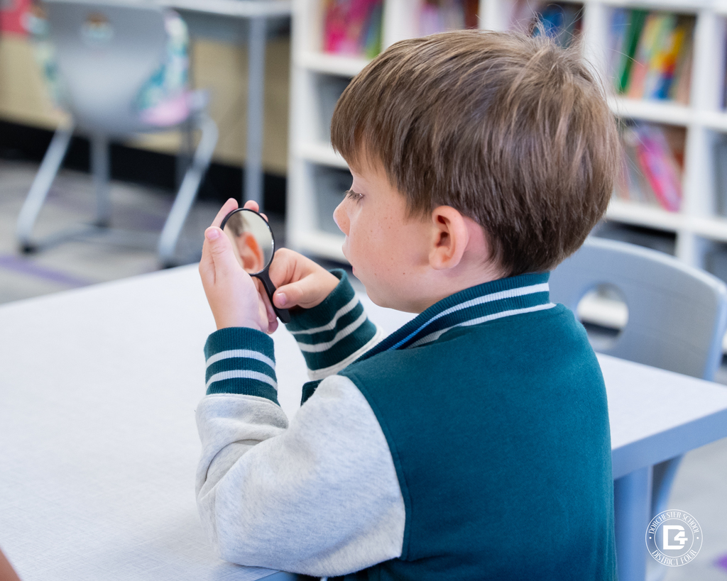 A student in a green and gray jacket holds a small mirror and studies how their mouth moves while saying the /k/ sound during class.