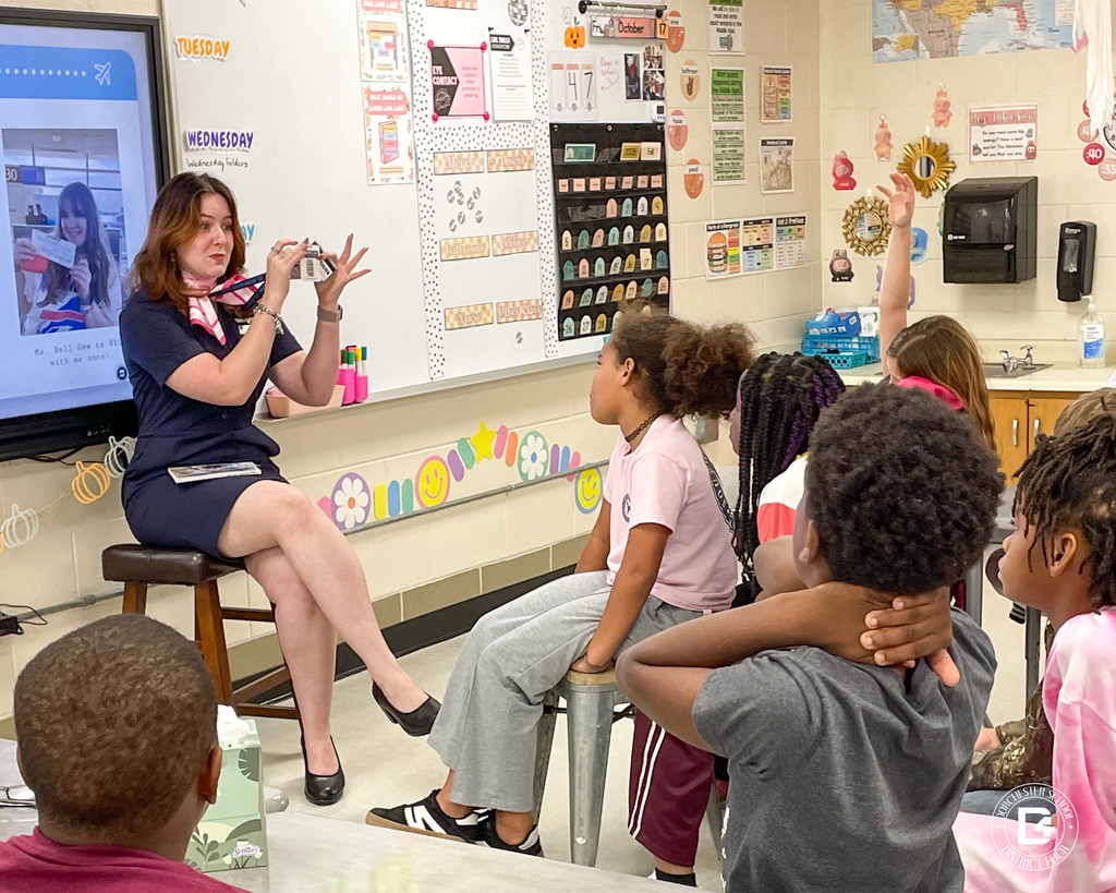 American Airlines flight attendant Tei Hurst sits on a stool at the front of a classroom, holding her lanyard and ID badge while speaking to attentive fourth-grade students. The students are seated and listening closely, with one raising a hand to ask a question. The classroom is decorated with educational posters and a world map.