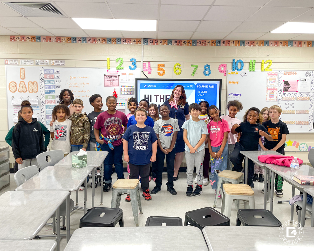 A group of fourth-grade students from Clay Hill Elementary stand together smiling inside their classroom with American Airlines flight attendant Tei Hurst. Ms. Hurst is centered behind the group, wearing her navy uniform and pink scarf. The classroom walls display colorful numbers, student work, and a large screen that reads “Hi There” with a boarding pass graphic.