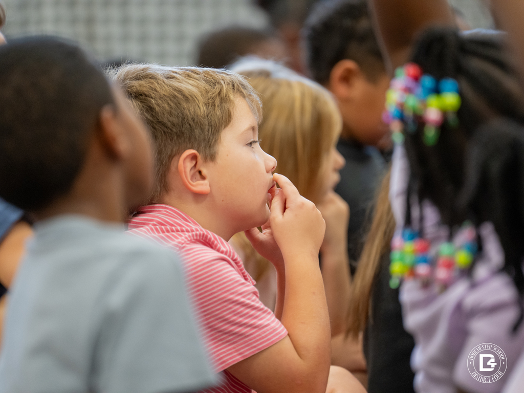 A young boy in a red-striped shirt sits cross-legged on the floor, resting his chin on his hand as he listens to the author’s story. Other students sit nearby, facing forward toward the speaker.