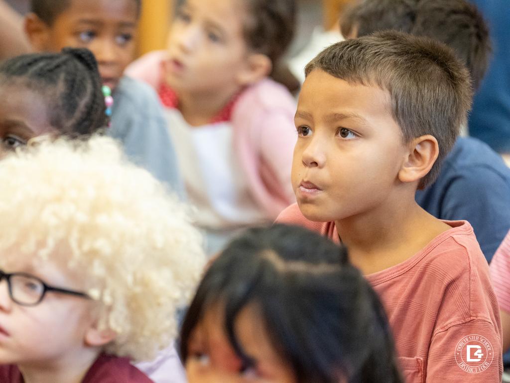 A young boy with short brown hair looks toward the front of the room with a focused expression. Around him, other students sit closely together and watch the presentation.