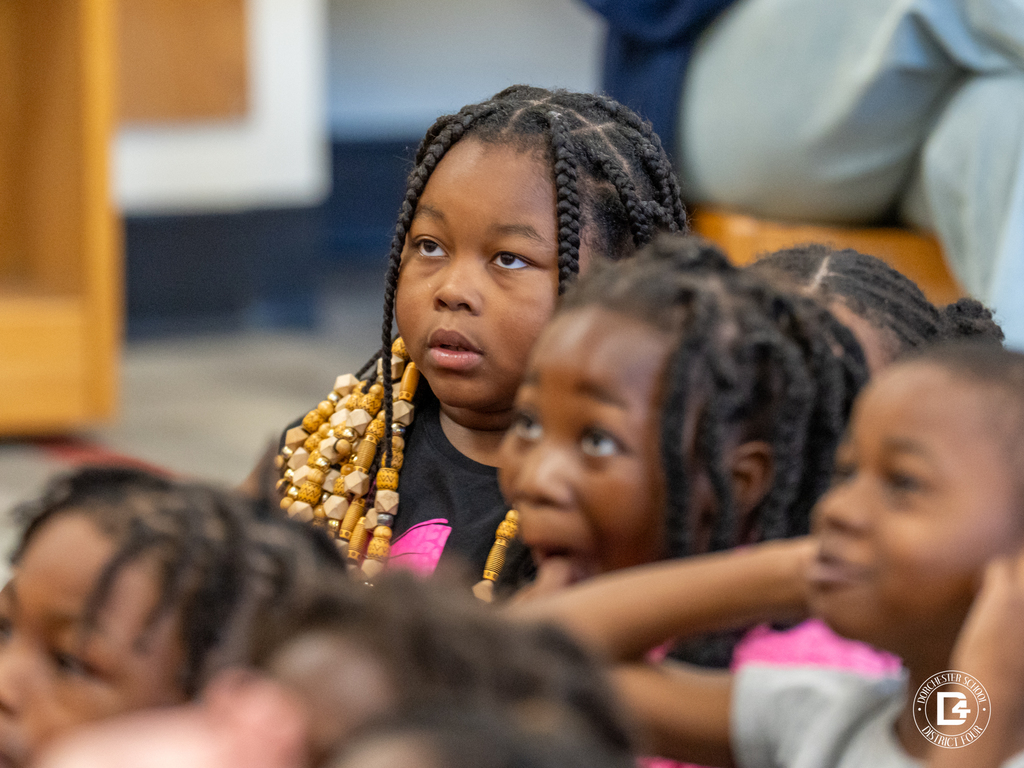 Several young students sit close together on the floor, looking toward the front of the room. A girl with long braids and gold beads gazes forward, appearing thoughtful and engaged.
