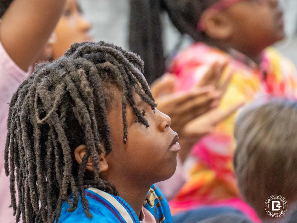 A young student with short dreadlocks looks upward attentively while listening to the author’s presentation. Other students and bright classroom colors appear slightly blurred in the background.