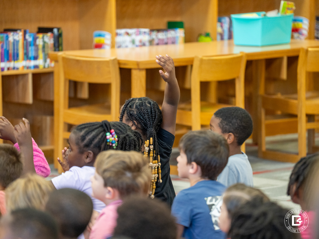 A group of elementary students sit on the floor of a library, listening attentively. One student with braided hair and colorful beads raises her hand to answer a question. Bookshelves and chairs line the background.