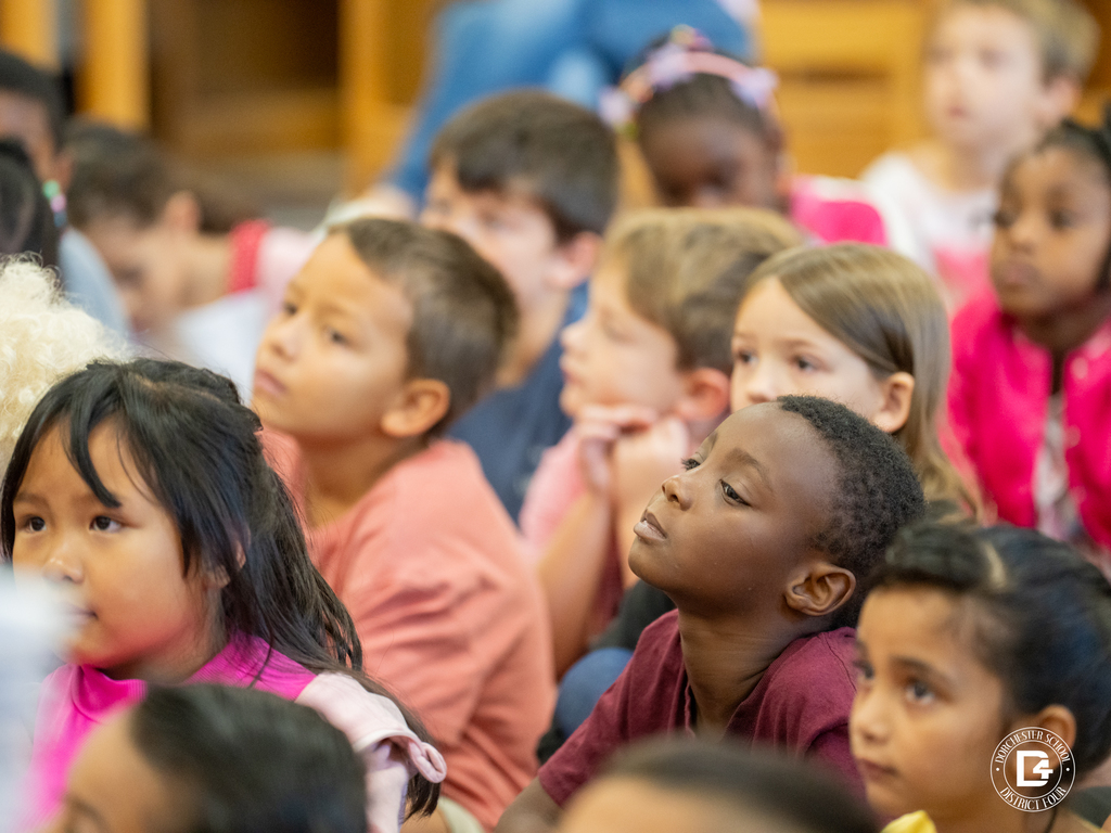A group of diverse elementary students sit quietly on the floor, watching the speaker at the front of the room. Their expressions show focus and curiosity.