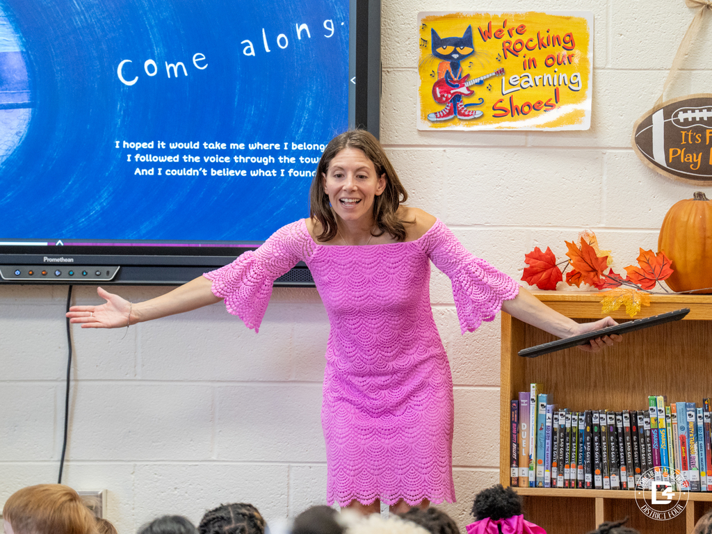 Author Gina Casazza stands at the front of a classroom, wearing a bright pink dress and holding a tablet. She gestures with open arms while speaking to a group of elementary students. Behind her, a large screen displays the words “Come along,” and the classroom is decorated with fall leaves and a Pete the Cat poster.