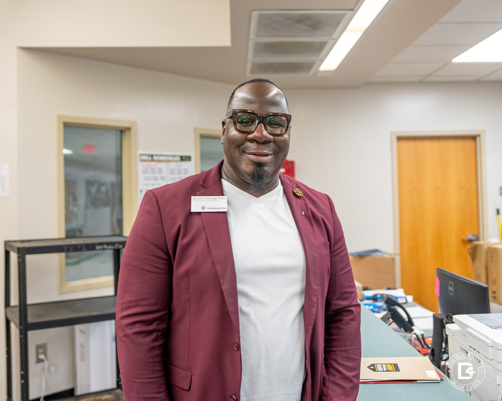 A staff member wearing glasses, a maroon blazer, and a name badge stands at a service counter, smiling in a school office area.