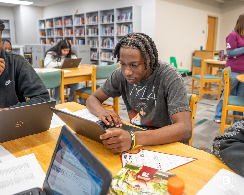 A student with short braids works on a laptop at a library table during College Application Day, with papers and other laptops on the table and bookshelves in the background.