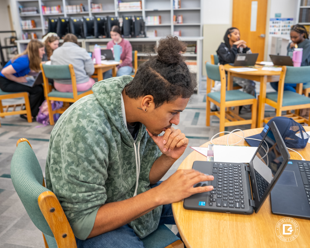 A student in a green camouflage hoodie leans over a laptop, concentrating at a library table while groups of students work at nearby tables.
