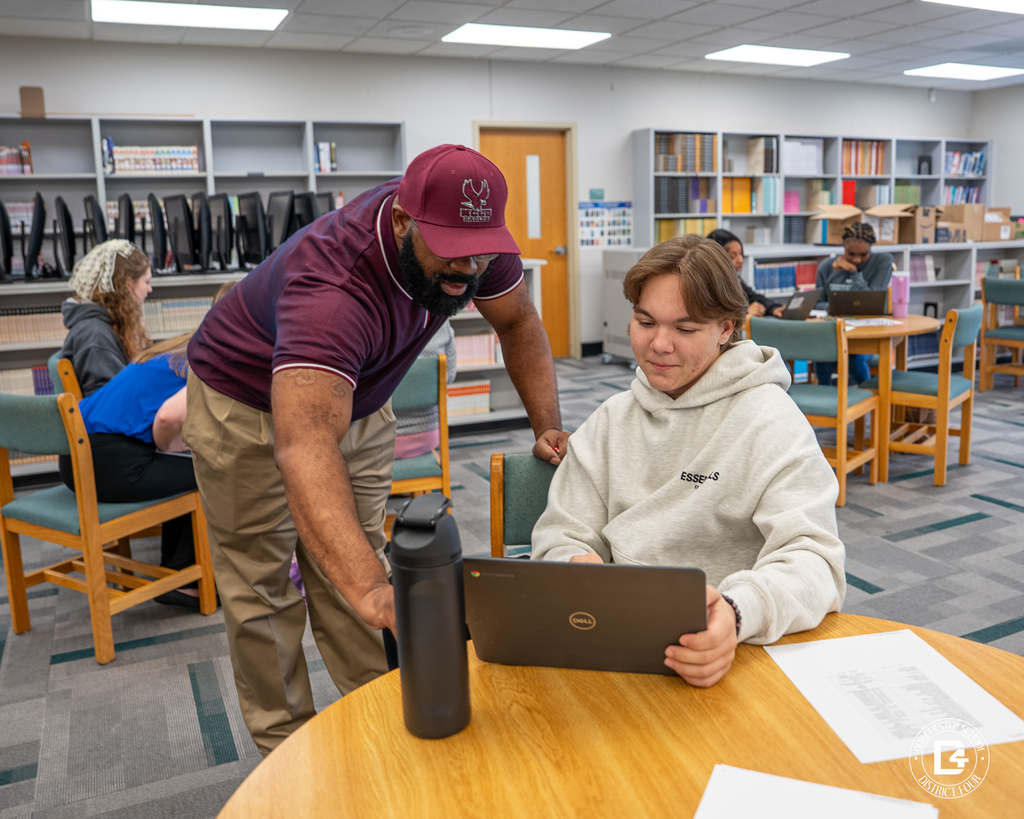 A staff member in a maroon polo bends to assist a student in a white hoodie who is viewing a laptop at a round table in the library.