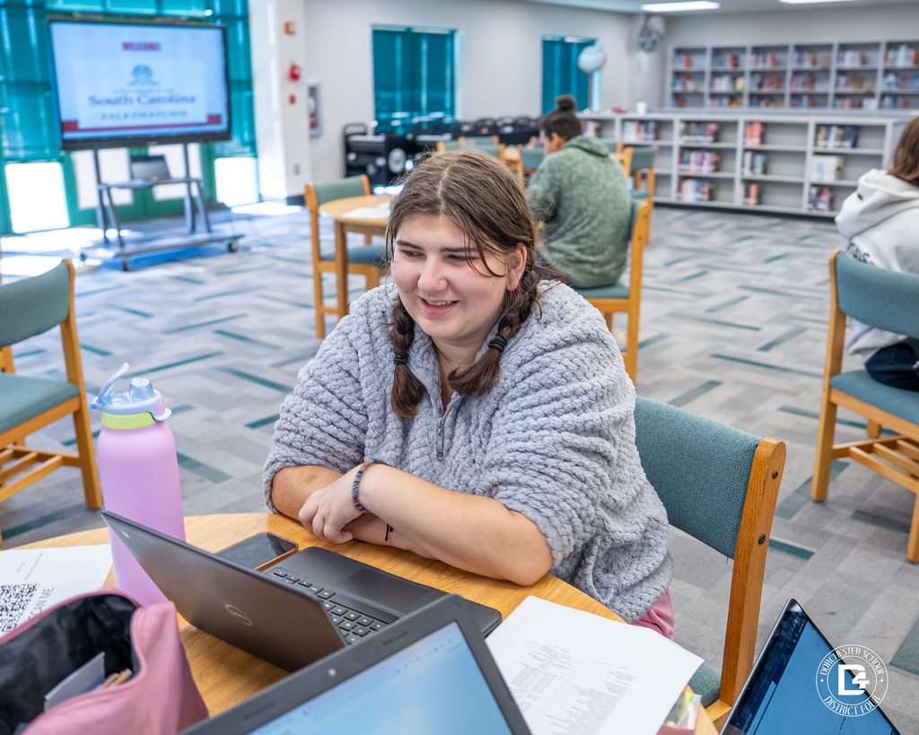 A student in a gray sweater smiles while working on a laptop in the library, with papers and a water bottle on the table and a presentation screen in the distance.