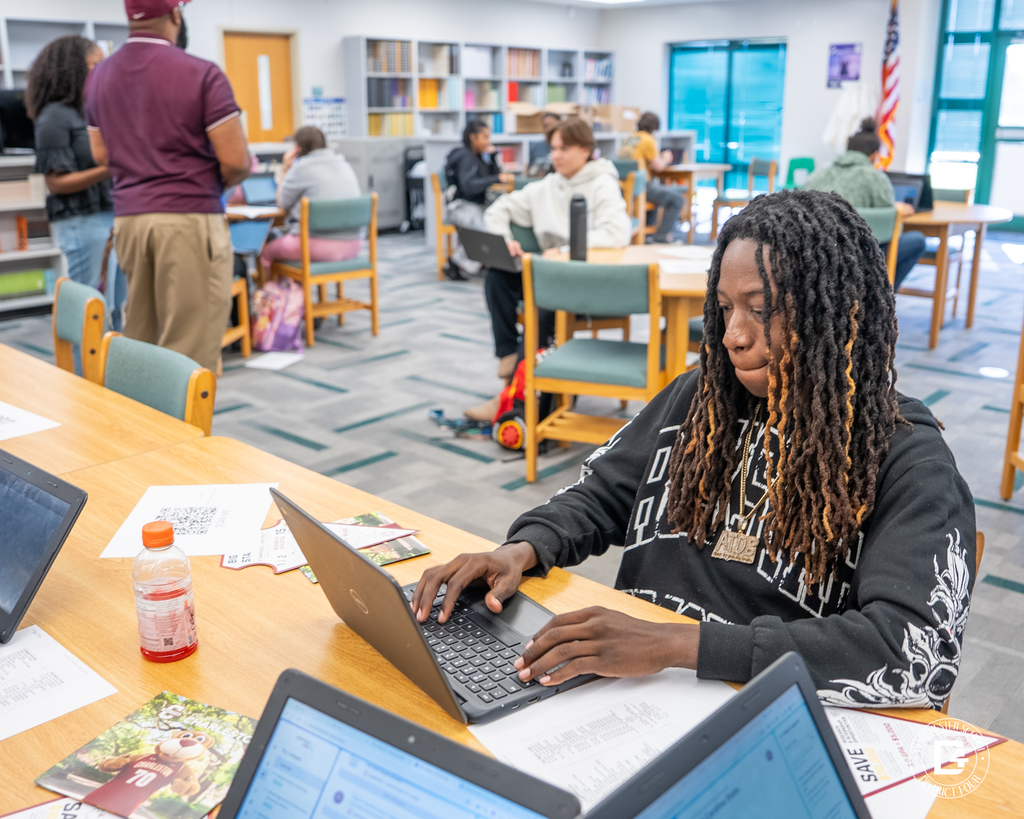 A student with long dreadlocks types on a laptop at a library table, with a drink bottle, flyers, and other students working in the background.