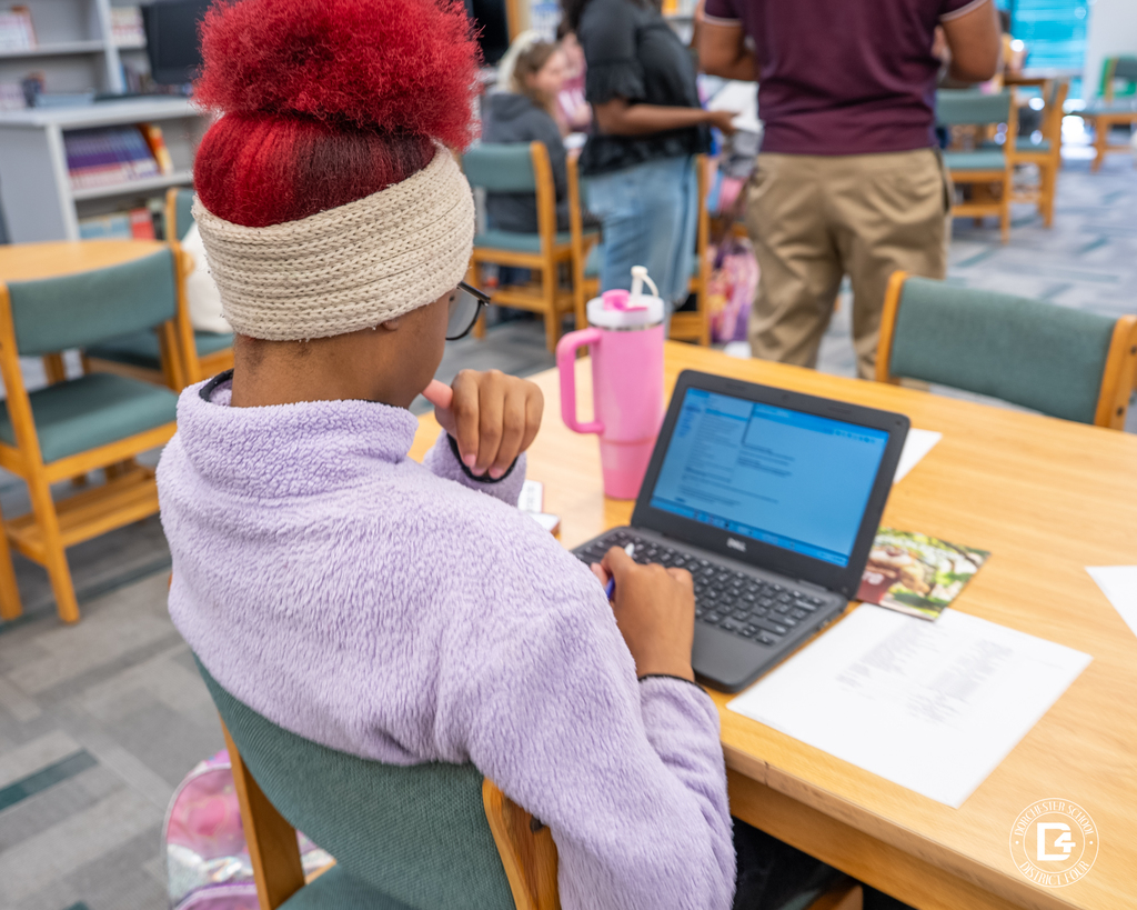 A student with bright red hair in a cream headband and lavender fleece works on a laptop at a library table, with a large pink water bottle nearby.