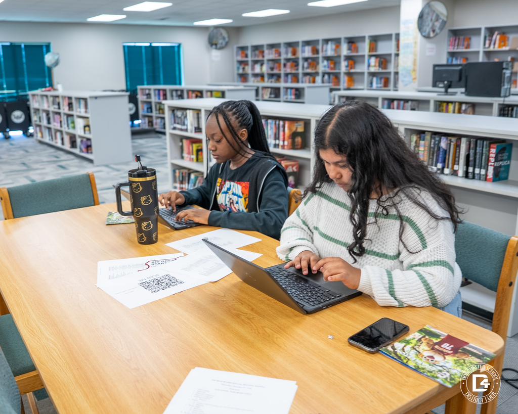 Two students sit side by side at a rectangular library table, each typing on a laptop, with papers, a phone, and a large tumbler on the table and bookshelves behind them.