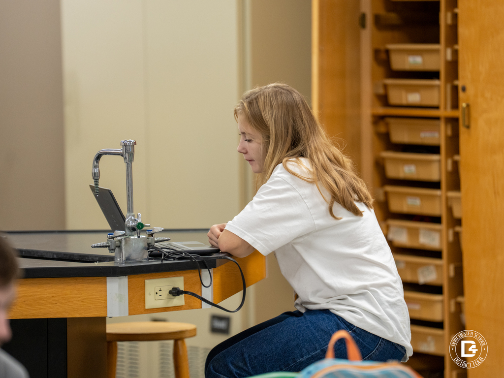 A student with long blonde hair sits on a stool at a lab table, working on a laptop connected to an outlet. Wooden storage cabinets are visible in the background.