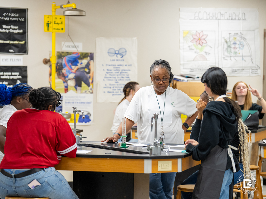 Mrs. Frazier, a teacher wearing a white shirt, stands with a group of students gathered around a black lab table. Students discuss their experiment as classroom safety posters and equipment are visible in the background.
