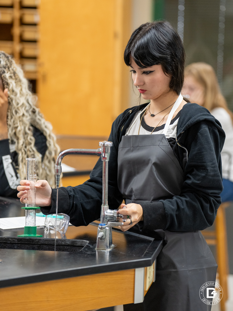 A student wearing a black hoodie and lab apron carefully fills a graduated cylinder with water at a science lab sink while focusing on the experiment.