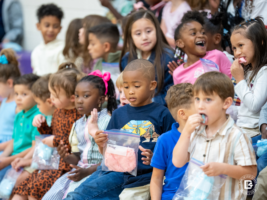 Young elementary students sit in a gym bleacher row, each holding a small bag of cotton candy. Some children are eating, while others smile and chat with friends during the reward event.