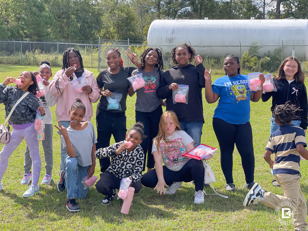 A group of students poses outdoors, smiling and holding bags of cotton candy. Some students are eating their treats while others make peace signs, celebrating their good attendance.