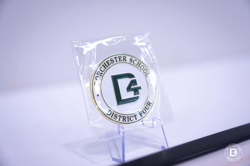 A close-up view of the Dorchester School District Four Superintendent’s Challenge Coin, gold-rimmed with the DD4 logo centered in green and white, displayed on a small clear stand.