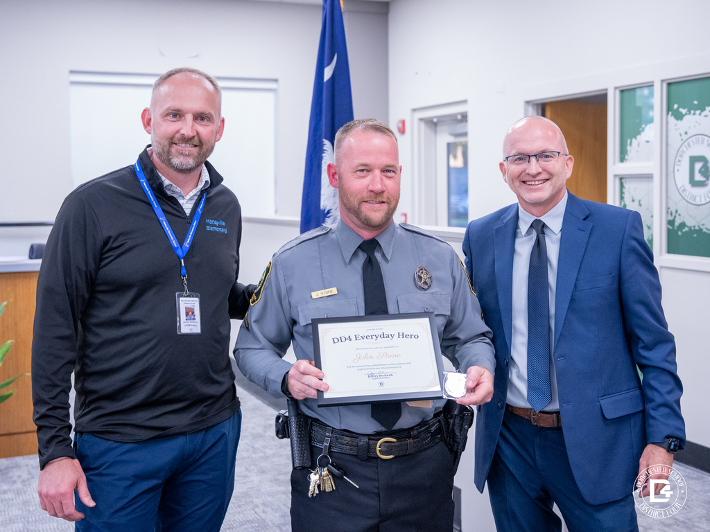 Officer John Stone, School Resource Officer at Harleyville Elementary, stands smiling with Principal Jeremy Wolfgang and Superintendent Jeffrey Beckwith after receiving his DD4 Everyday Hero award and Challenge Coin.