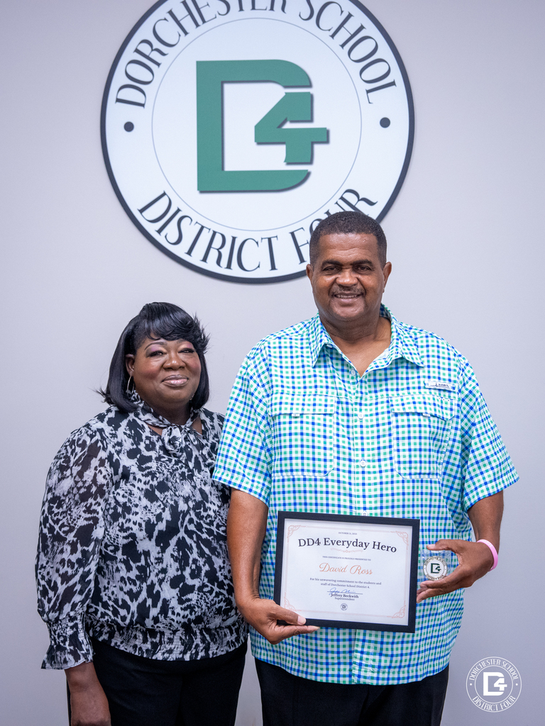 David Ross stands in front of the Dorchester School District Four logo, holding his DD4 Everyday Hero certificate and Challenge Coin alongside his wife smiling beside him.