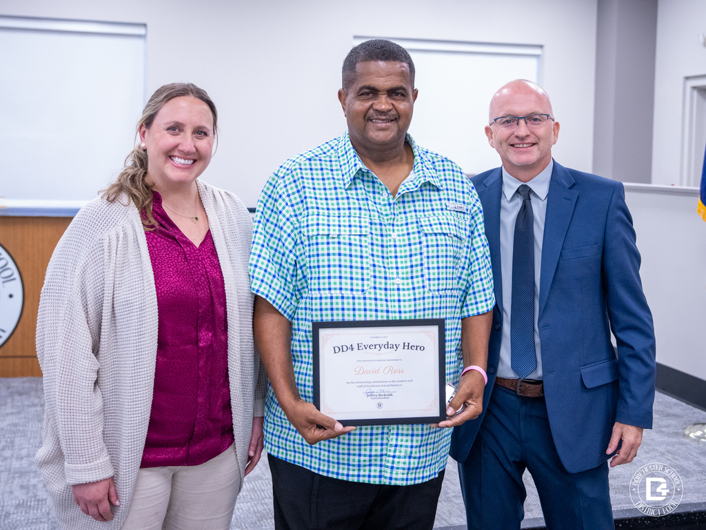 David Ross, DD4 Activity Bus Driver, holds his DD4 Everyday Hero certificate and Challenge Coin while standing with Assistant Athletic Director Wylie McCall and Superintendent Jeffrey Beckwith.