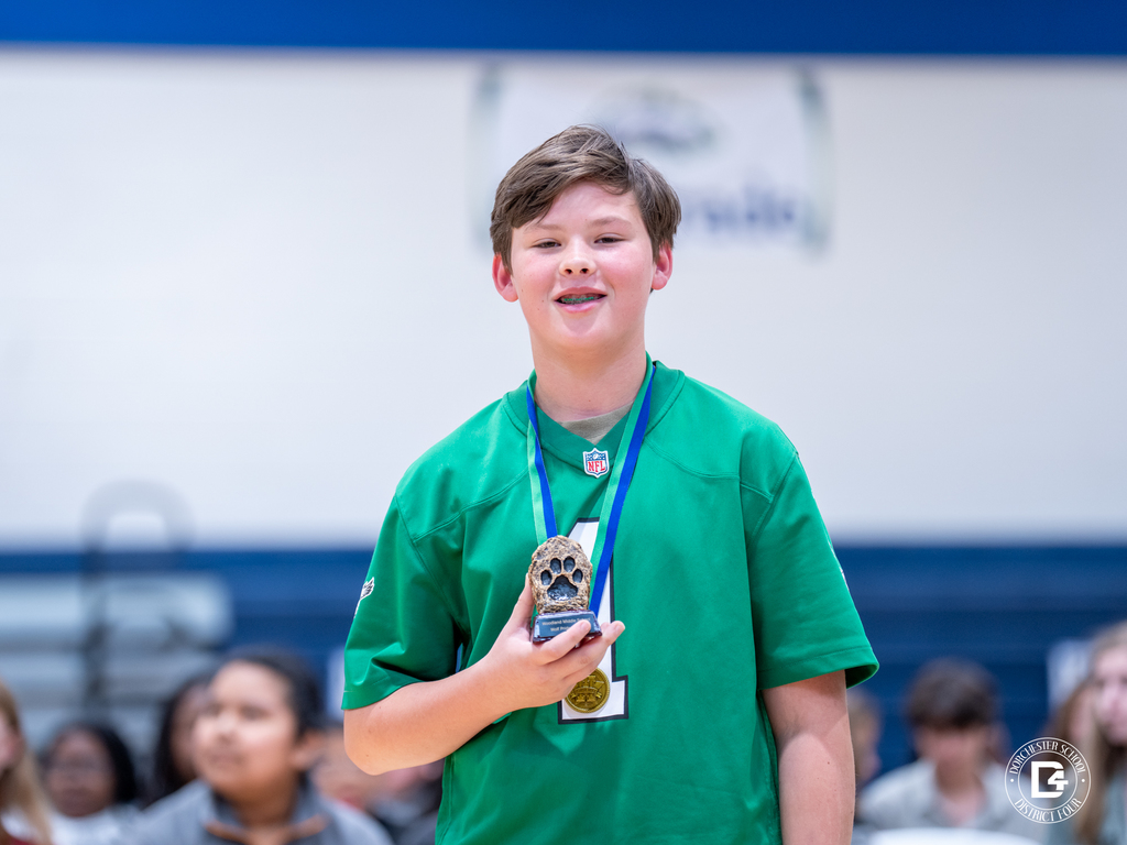 A student wearing a green shirt smiles while holding the Woodland Middle School Wolf Pride Award trophy and gold medal.