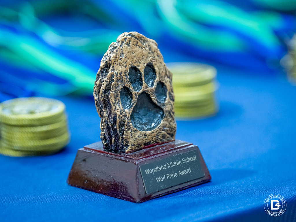 A close-up of the Woodland Middle School “Wolf Pride Award” trophy featuring a paw print design sits on a blue table surrounded by medals.