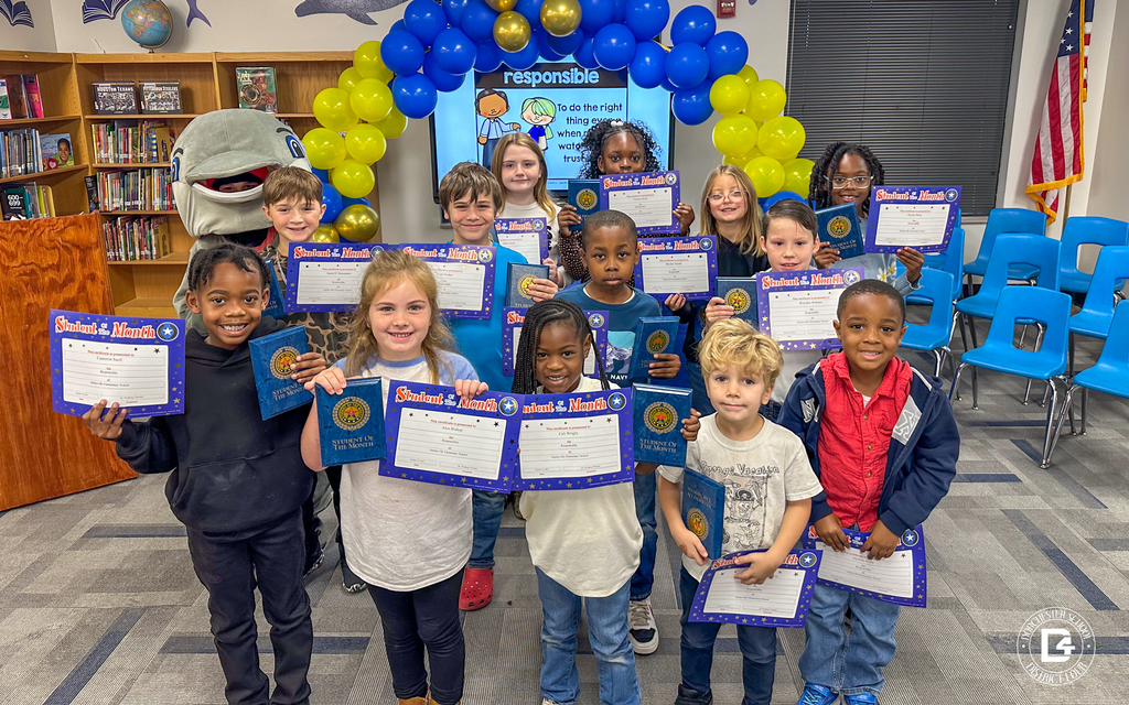 A group of Harleyville Elementary School students smile proudly while holding their “Student of the Month” certificates and plaques. Behind them, the school’s dolphin mascot stands near a display decorated with blue and yellow balloons and a screen showing the word “Responsible.” The celebration takes place in a bright library with bookshelves and blue chairs in the background, honoring students recognized for demonstrating responsibility.
