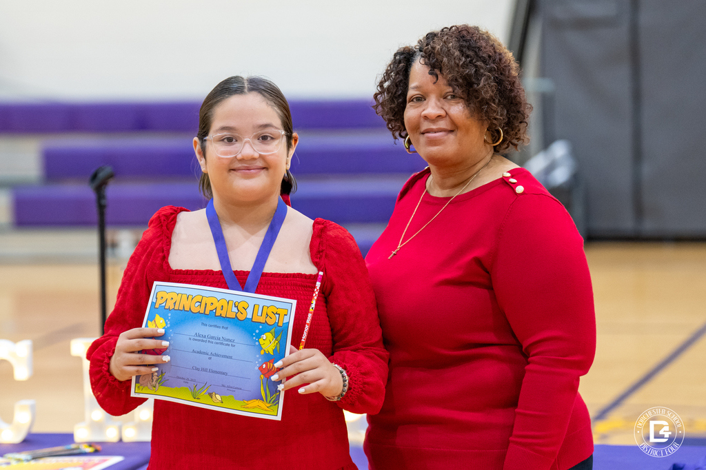 A Clay Hill Elementary student smiles while holding a “Principal’s List” certificate and wearing a medal beside a staff member in a red sweater. The ceremony takes place in the school gym, with purple bleachers and a table of awards in the background.