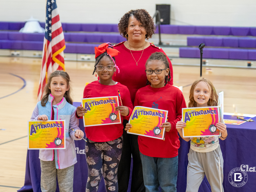 Four Clay Hill Elementary students stand in front of a staff member, each holding a bright “Perfect Attendance” certificate. They smile proudly in the school gym, with the American flag and purple bleachers behind them.