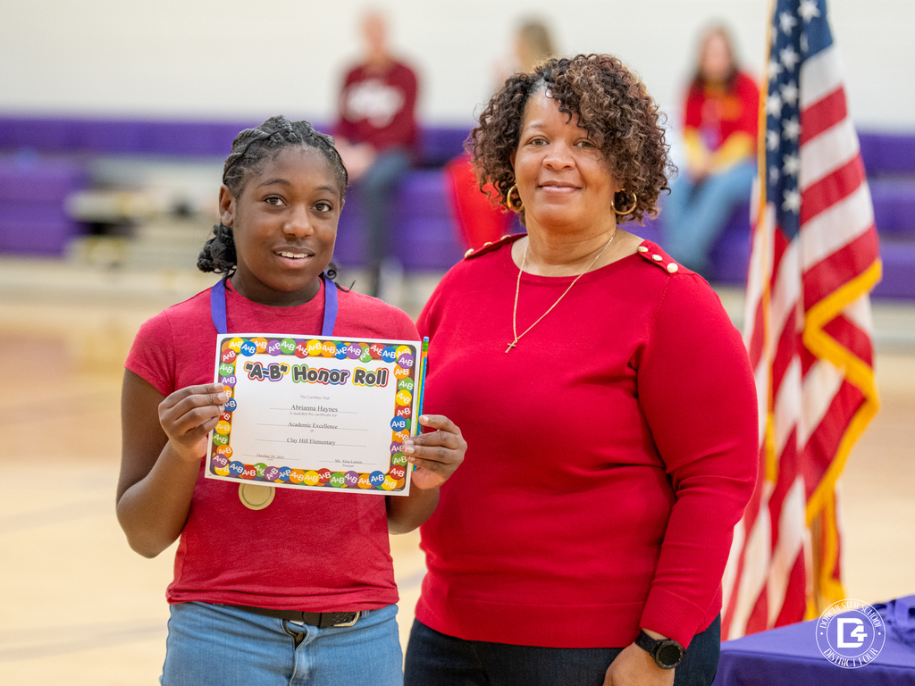 A Clay Hill Elementary student proudly holds an “A/B Honor Roll” certificate beside a staff member in a red sweater. They stand in front of an American flag and purple bleachers during the school’s awards ceremony.