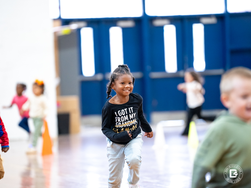 A young Harleyville Elementary student runs inside the gym with a joyful smile during the Pumpkin Run. Other children can be seen running in the background, and the gym doors and cones mark the running course.