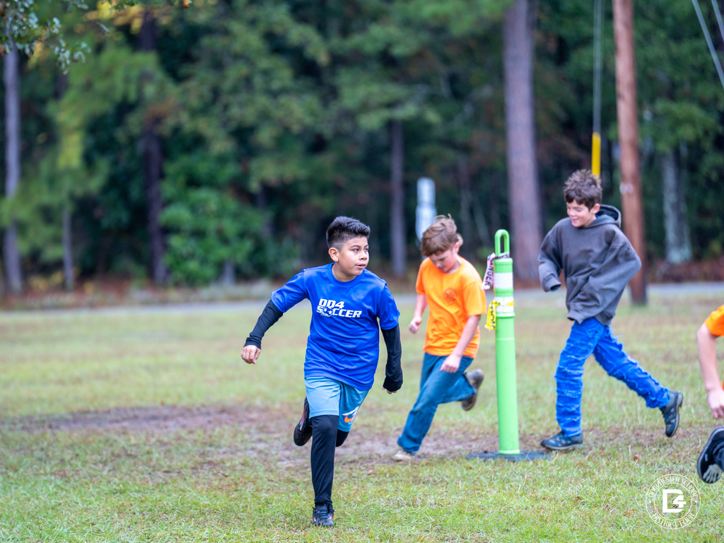 Fifth-grade students run outdoors during Harleyville Elementary’s Pumpkin Run. The ground is damp, and the runners, dressed in cool-weather clothes, sprint across the grass as trees and utility poles line the background.