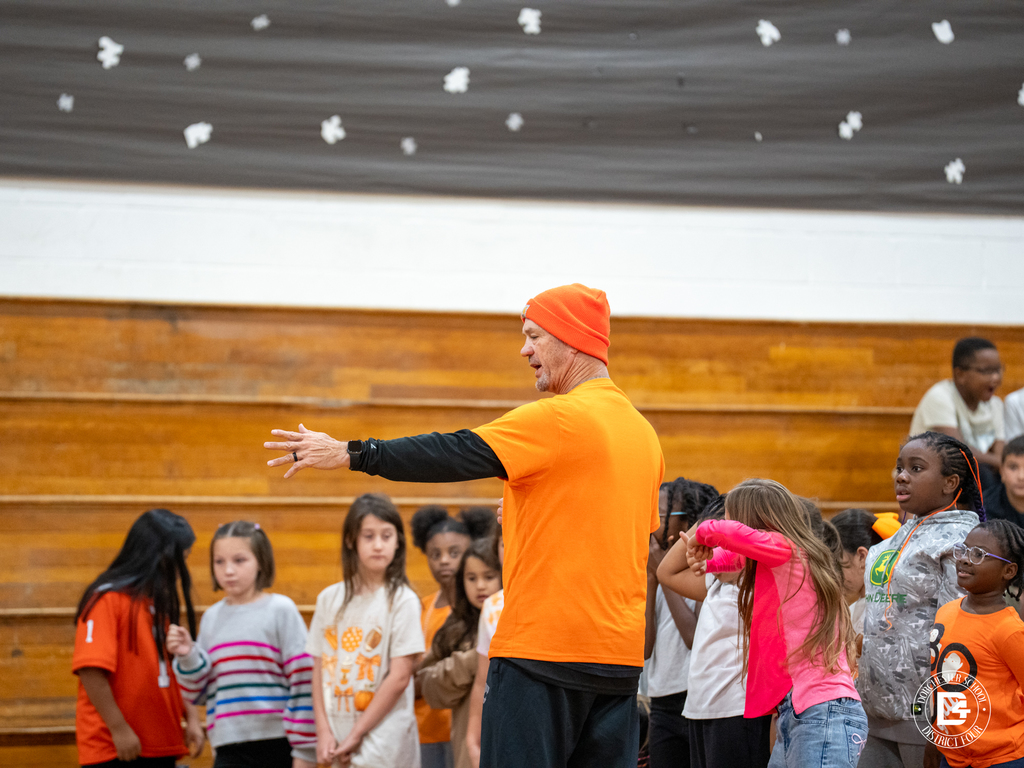 Physical Education teacher Stephen Jackson, dressed in orange with a matching beanie, gives directions to a group of Harleyville Elementary students gathered in the gym for the Pumpkin Run. Students listen attentively, wearing bright fall colors.